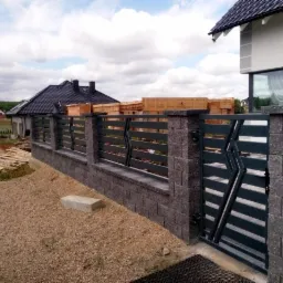 Nowoczesne ogrodzenie panelowe z bramą, słupki z szarego kamienia łupanego, poziome przęsła w kolorze antracytowym, na tle nowego domu w budowie i zieleni.