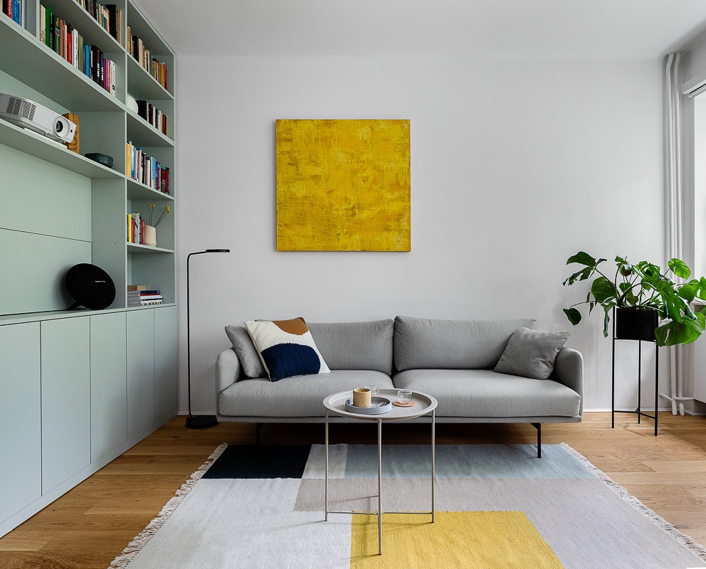Nowoczesny salon z szarą sofą, abstrakcyjnym żółtym obrazem na ścianie, miętową zabudową meblową z projektorem i głośnikiem oraz geometrycznym dywanem.