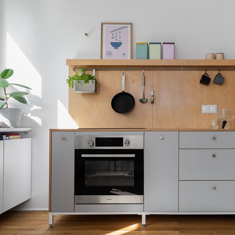 Jasna kuchnia z szarymi szafkami, drewnianym blatem i panelem ściennym z płyty OSB, na którym wiszą czarne naczynia i doniczka z ziołami, a na górnej półce stoją kolorowe puszki.