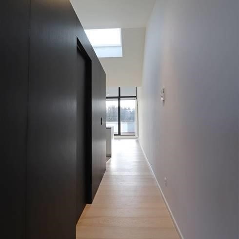 Wąski korytarz w nowoczesnym stylu z jasną drewnianą podłogą, ciemnymi, minimalistycznymi szafami po lewej stronie i białymi ścianami po prawej, zakończony oknem z widokiem na zieleń.
