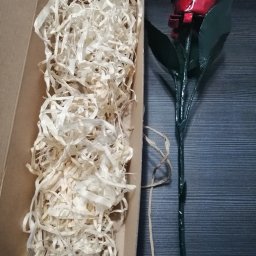 NITREX Rafał Nita - Ręcznie robiona metalowa róża, czerwona główka i zielona łodyga, leży obok pudełka wypełnionego wiórkami papierowymi.