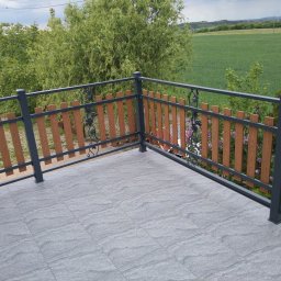NITREX Rafał Nita - Balkon z szarą, kamienną podłogą i balustradą z drewnianymi szczeblami oraz metalowymi, ozdobnymi elementami, widok na zielone pola i drzewa.