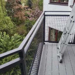 Czarna, metalowa balustrada balkonowa z siatkowym wypełnieniem i drabina oparta o ścianę budynku, widok na zielony ogród z kwitnącymi drzewami.