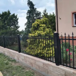 Czarny metalowy płot z dekoracyjnymi grotami, osadzony na niskim murku z kamienia elewacyjnego, na tle zieleni i budynku z oknem.