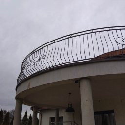 NITREX Rafał Nita - Kuta balustrada balkonowa z dekoracyjnymi elementami w kształcie liści na zaokrąglonym balkonie, widok z dołu na tle pochmurnego nieba.