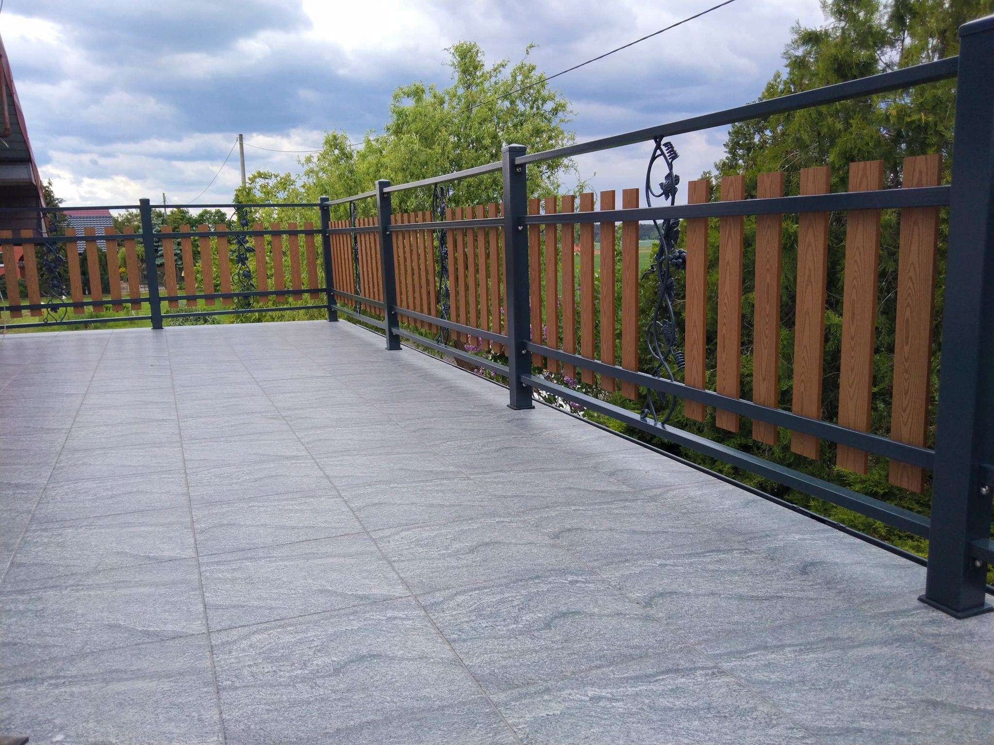 Balkon z szarą terakotą i balustradą z drewnopodobnych paneli pionowych oraz metalowymi elementami dekoracyjnymi z motywem roślinnym.