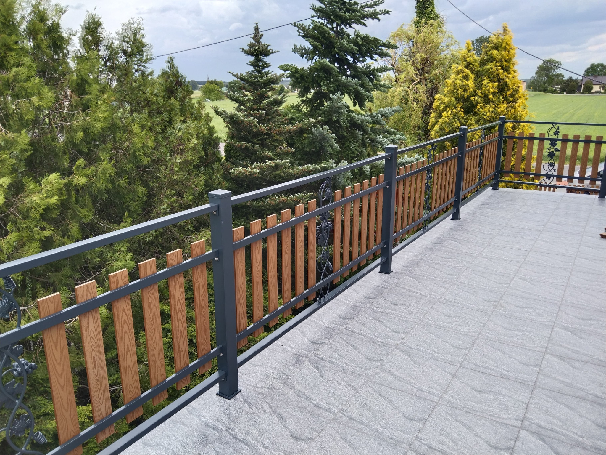 Szara metalowa balustrada z drewnianymi wypełnieniami na tarasie z szarych płytek, widok na zielone drzewa i pole w tle.