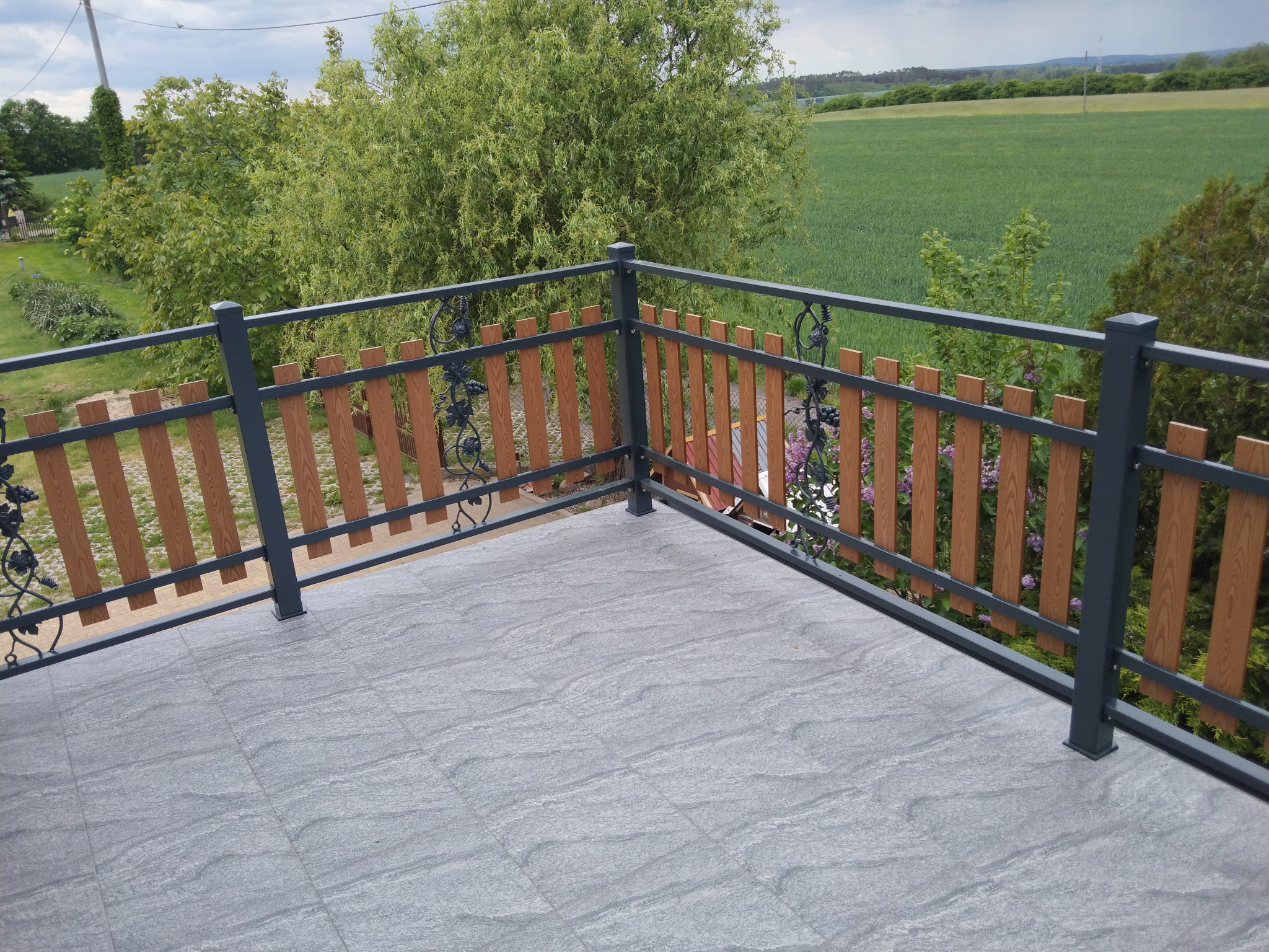 Balkon z szarą, kamienną podłogą i balustradą z drewnianymi szczeblami oraz metalowymi, ozdobnymi elementami, widok na zielone pola i drzewa.