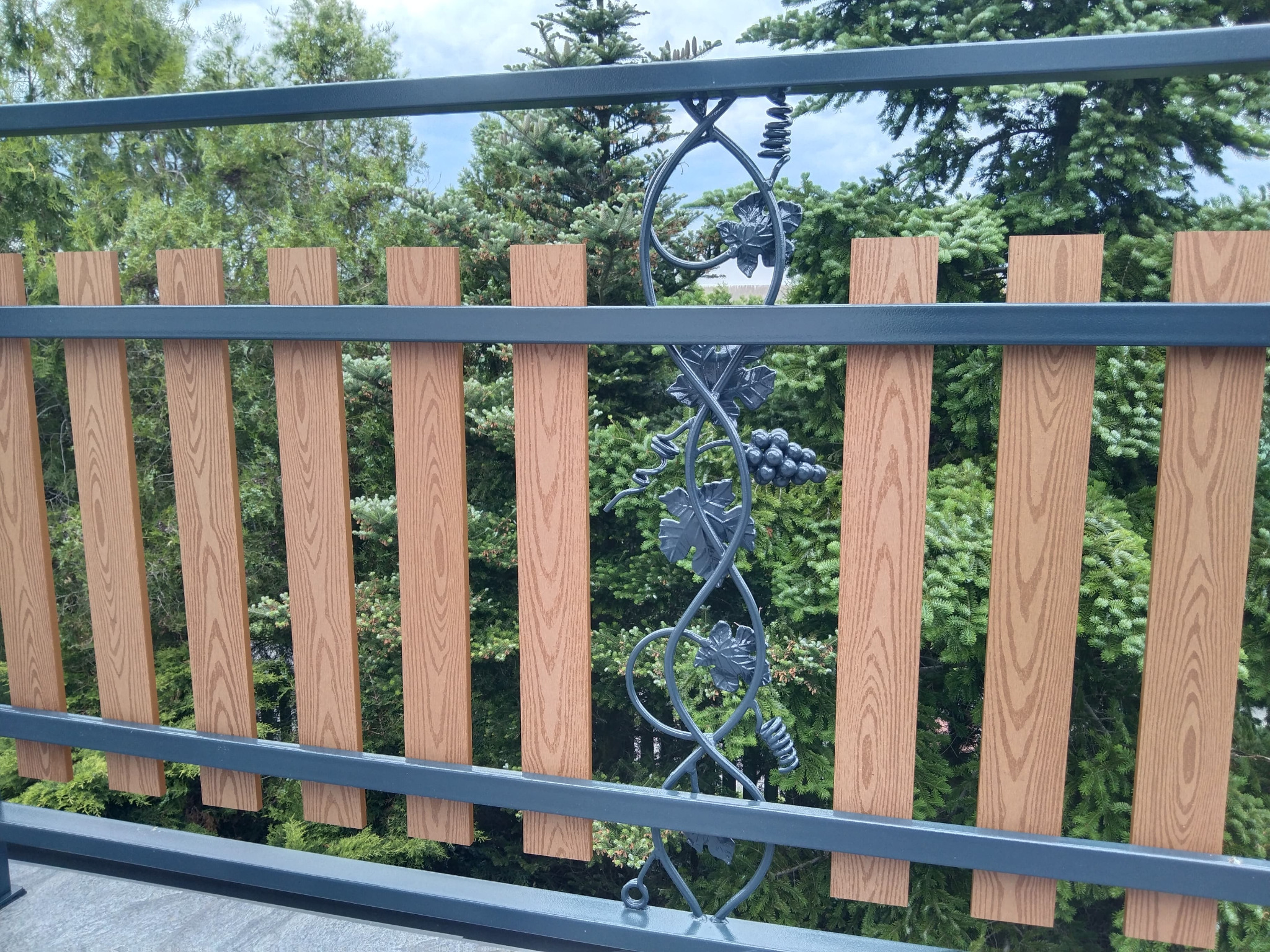 Szczegółowe ujęcie fragmentu ogrodzenia z drewnopodobnych paneli, z metalową, dekoracyjną aplikacją przedstawiającą winogrona i liście, na tle zielonych drzew.