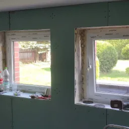 Montaż płyt kartonowo-gipsowych na ścianach wokół dwóch okien Aluplast, widoczne narzędzia, butelka Coca-Coli na parapecie, surowy stan wykończenia sufitu.