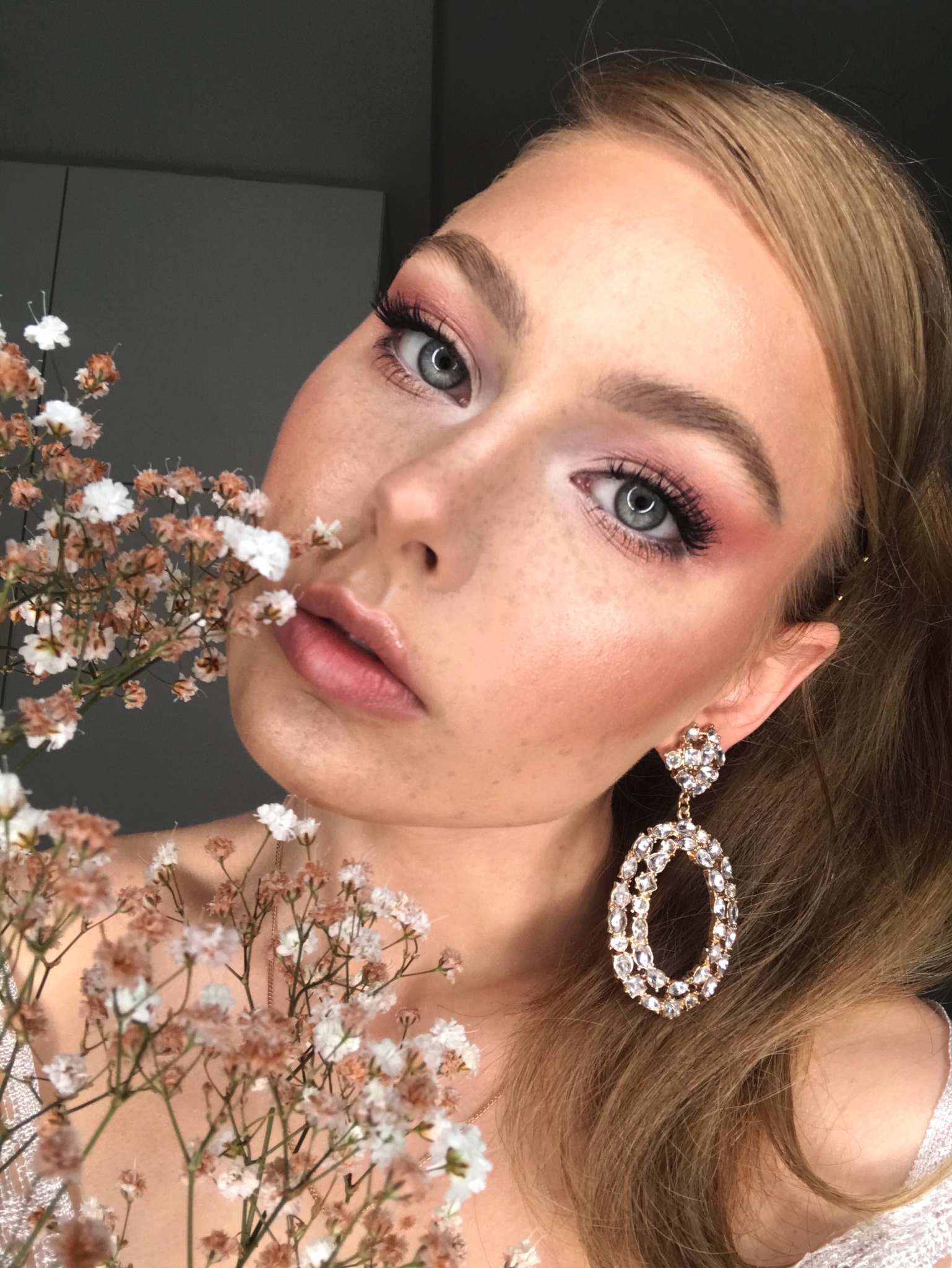 Portret kobiety z delikatnym makijażem w odcieniach różu, długimi rzęsami i błyszczącymi ustami, pozującej z gałązką gipsówki i dużym, ozdobnym kolczykiem.