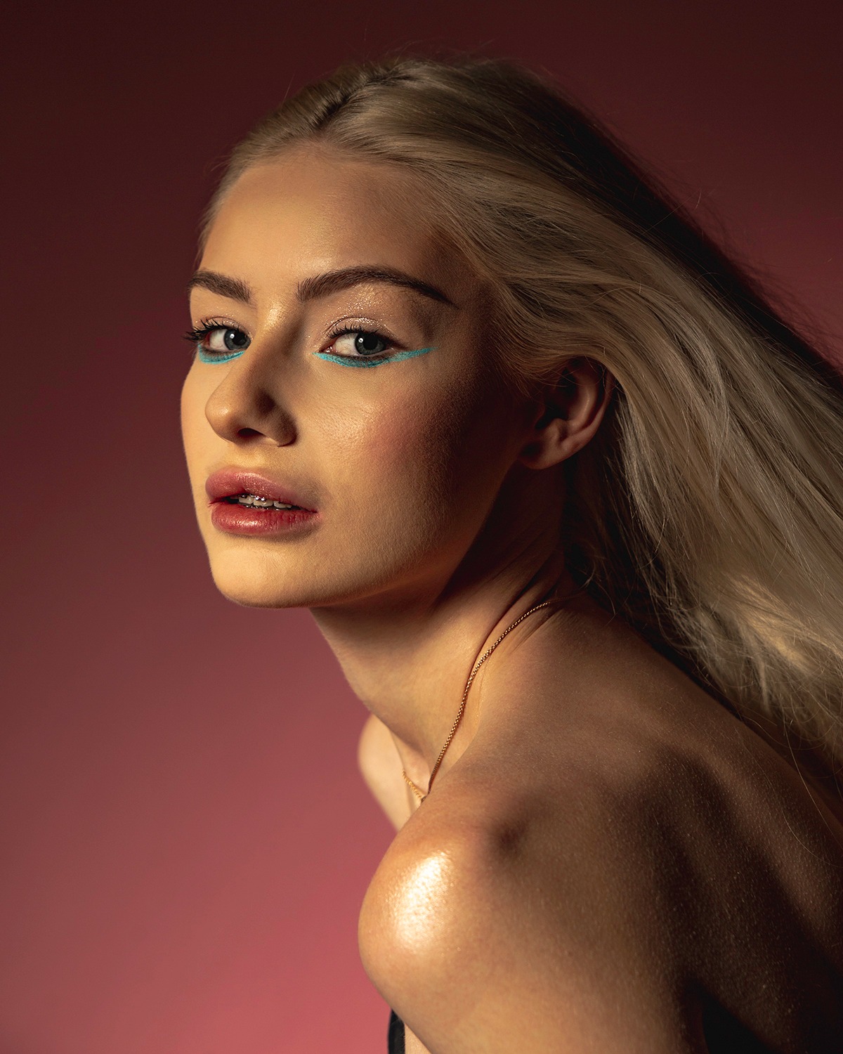 Portret kobiety z platynowymi włosami i turkusowym eyelinerem, spoglądającej przez ramię na tle różowego studia.