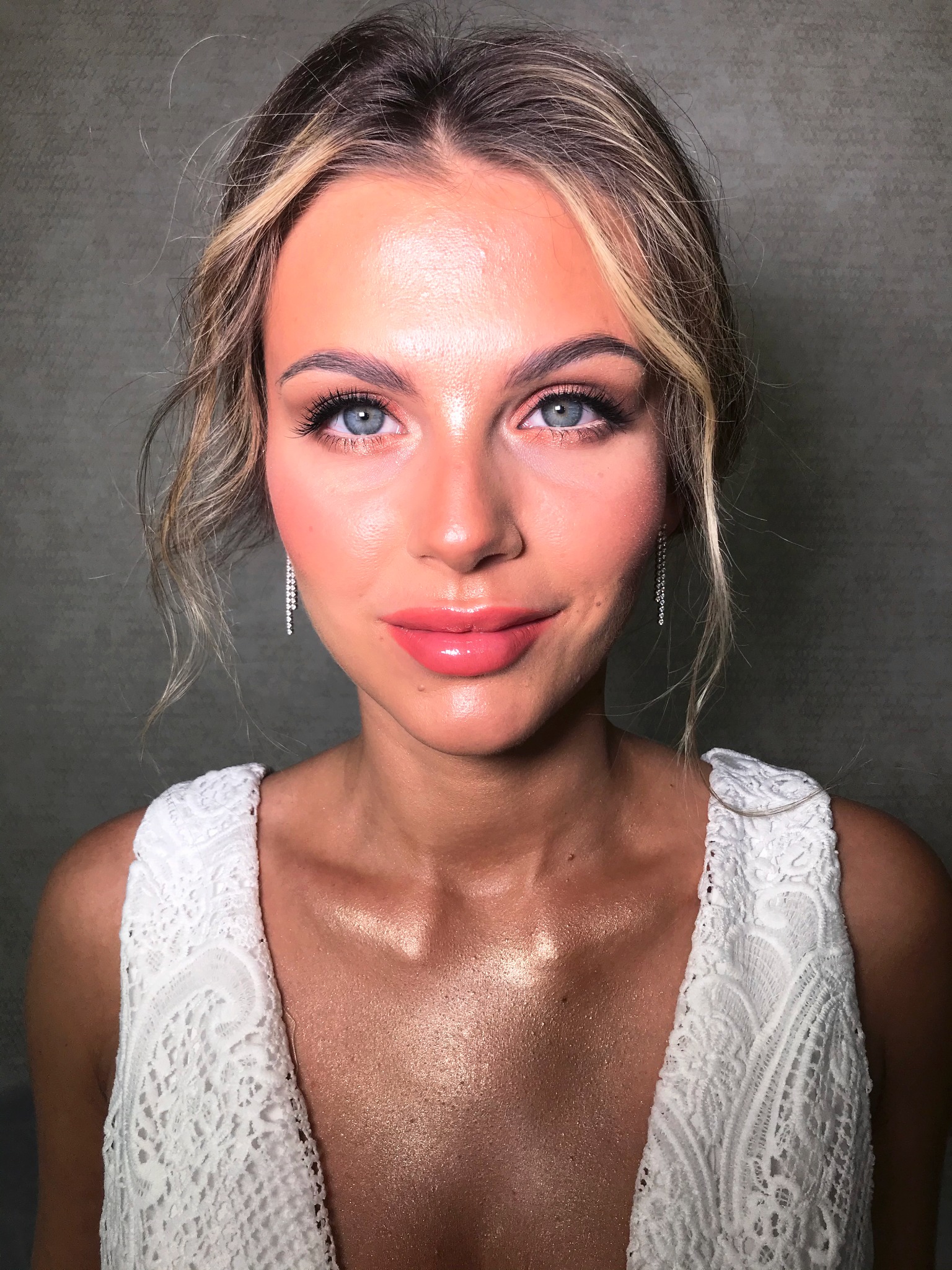 Portret kobiety z blond włosami upiętymi w kok, z delikatnym, rozświetlonym makijażem, długimi rzęsami i błyszczącymi ustami, ubrana w białą koronkową sukienkę.