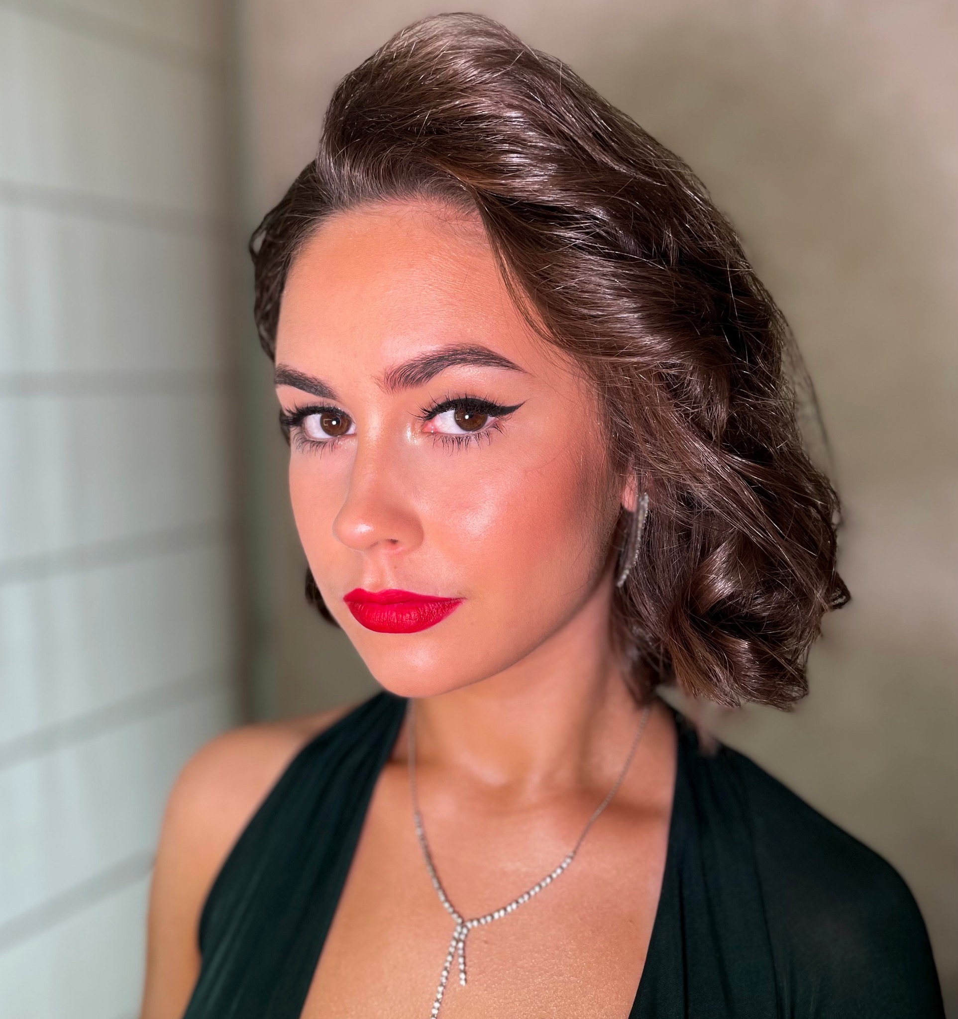 Portret kobiety z mocnym makijażem: czerwone usta, czarny eyeliner, podkreślone kości policzkowe, elegancka fryzura i biżuteria.