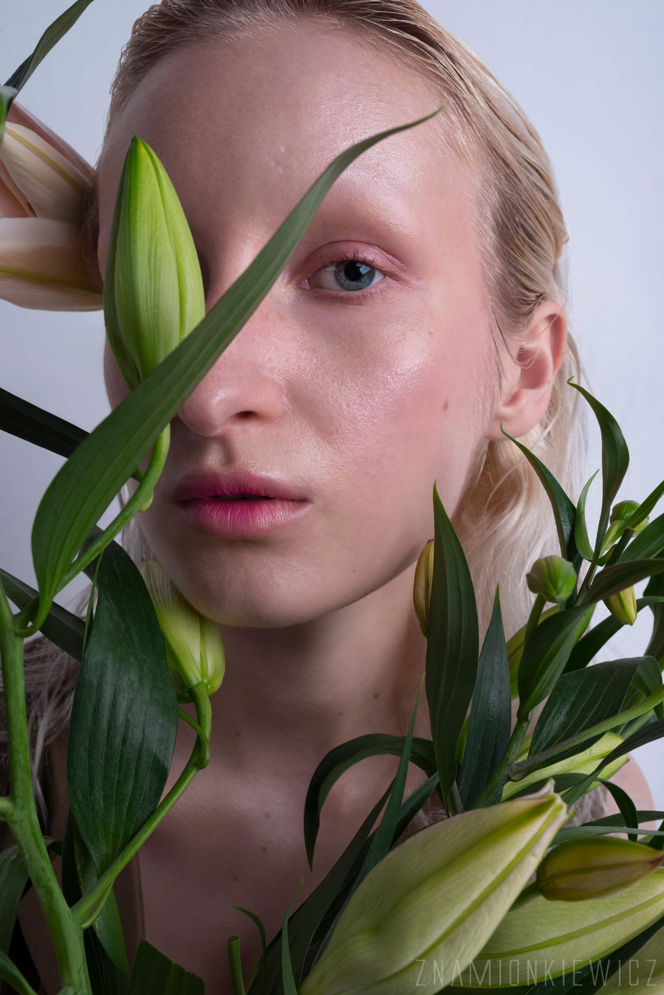 Portret kobiety z delikatnym, różowym rumieńcem i błyszczącymi ustami, otoczonej zielonymi liliami, z jednym okiem wyraźnie widocznym.