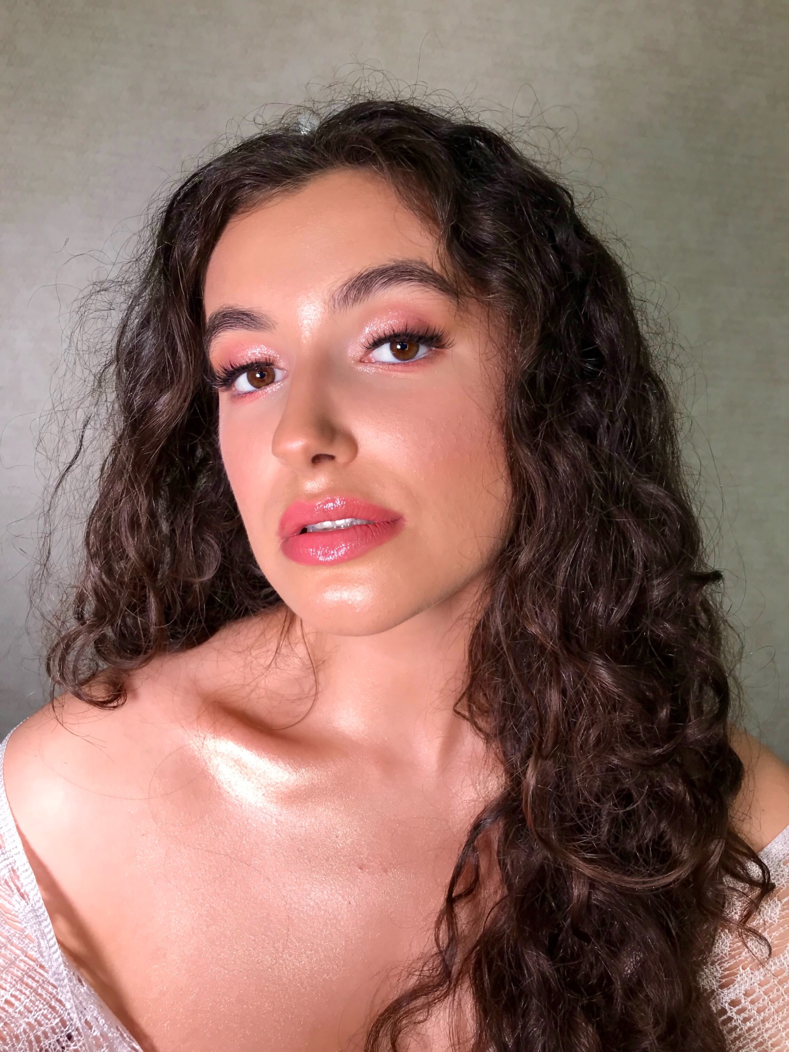 Portret kobiety z błyszczącym, różowym makijażem oczu i ust, podkreślonymi brązowymi oczami i burzą ciemnych, kręconych włosów, w delikatnej, białej bluzce z koronki.