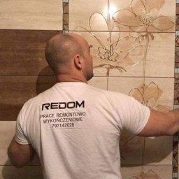 Redom - Pracownik w koszulce z logo firmy REDOM, układający beżowe płytki z motywem kwiatowym w łazience.