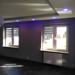 Redom - Szare wnętrze pokoju po remoncie z dwoma oknami z żaluzjami, grzejnikiem pod oknem, ozdobnym oświetleniem sufitowym LED w kolorze niebieskim i lampą z kryształkami