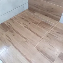 Ułożona podłoga z płytek imitujących drewno w łazience, z widocznym odpływem liniowym w rogu i szarą płytką ścienną u góry.