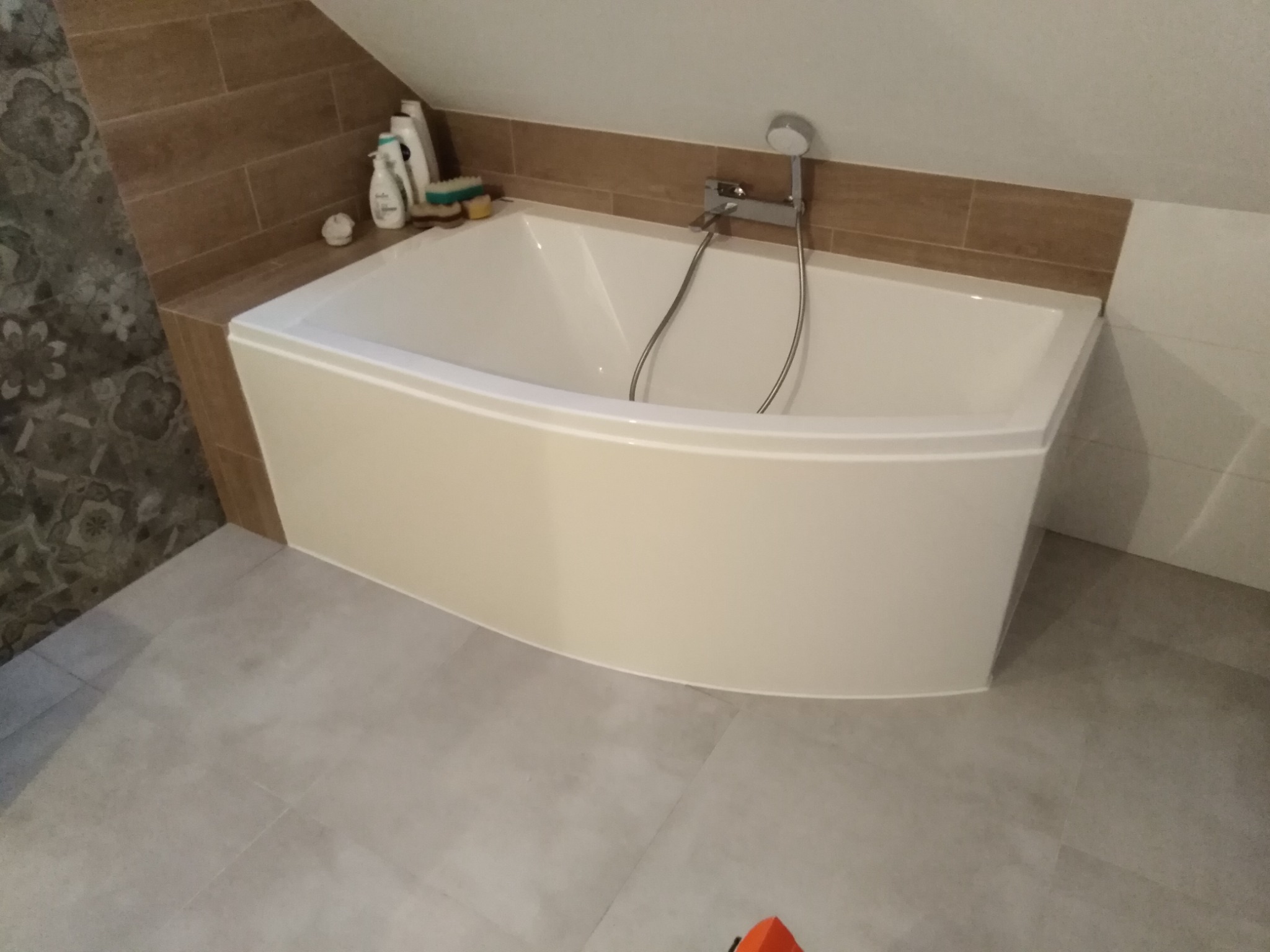 Biała, narożna wanna w łazience z beżowymi i szarymi płytkami, widok z góry, na wannie kosmetyki i bateria prysznicowa.