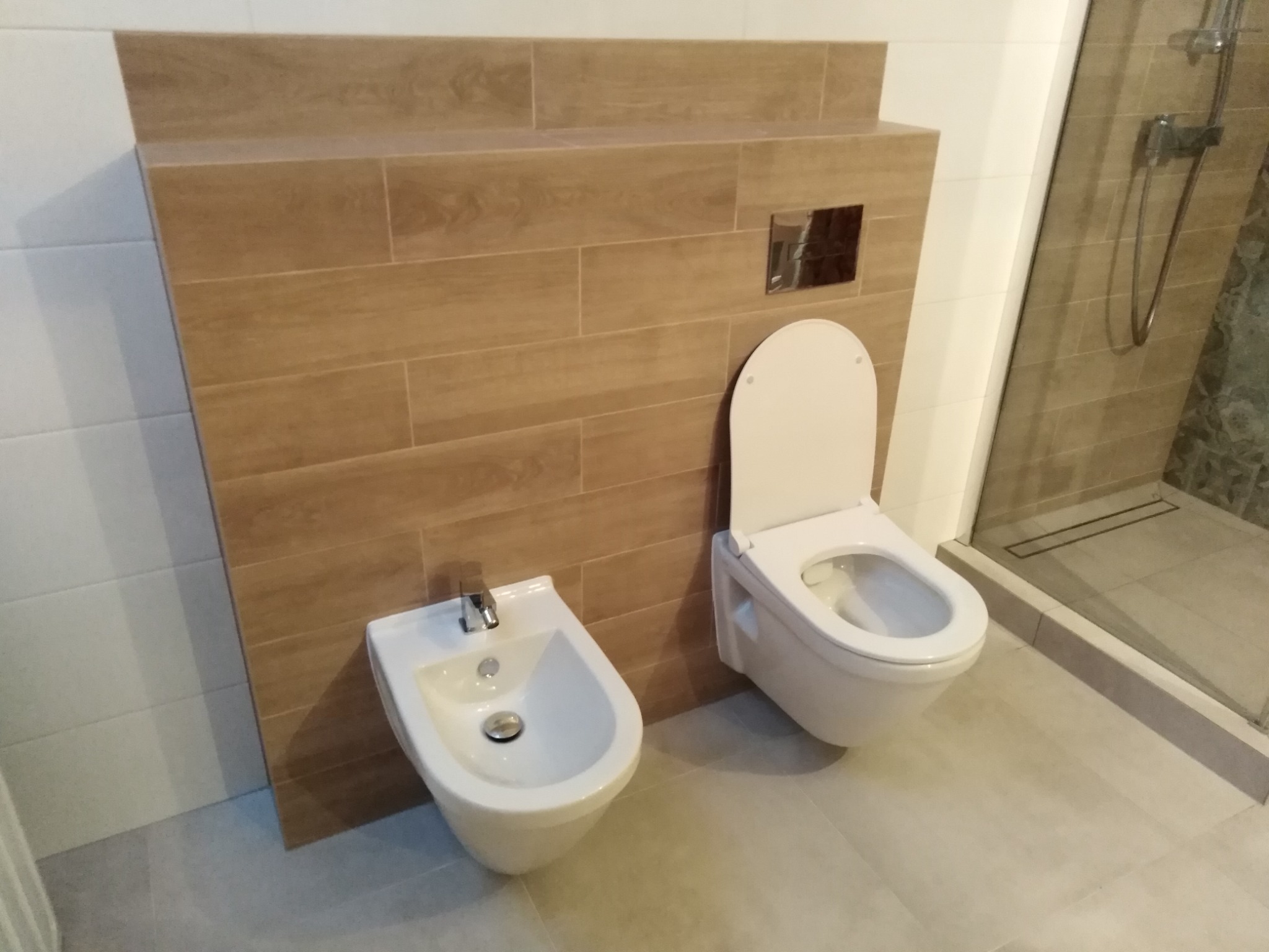 Nowoczesna łazienka z podwieszaną toaletą i bidetem na tle ściany wyłożonej płytkami imitującymi drewno, widoczna kabina prysznicowa z odpływem liniowym.