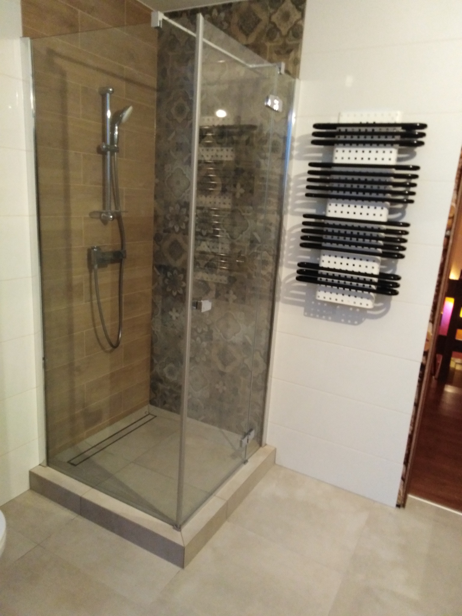 Szklana kabina prysznicowa z odpływem liniowym, brązowe płytki imitujące drewno na jednej ścianie i wzorzyste płytki na drugiej, grzejnik drabinkowy na białej ścianie.