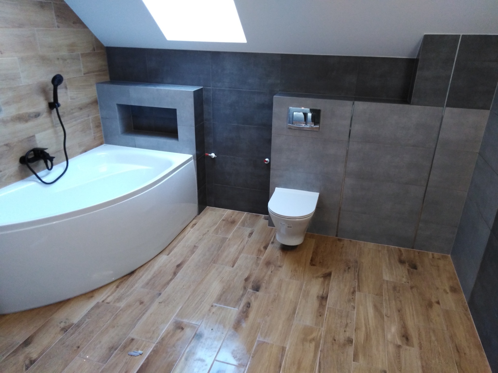 Nowoczesna łazienka na poddaszu z drewnopodobną podłogą, asymetryczną wanną i szarą glazurą, oświetlona światłem wpadającym przez okno dachowe.