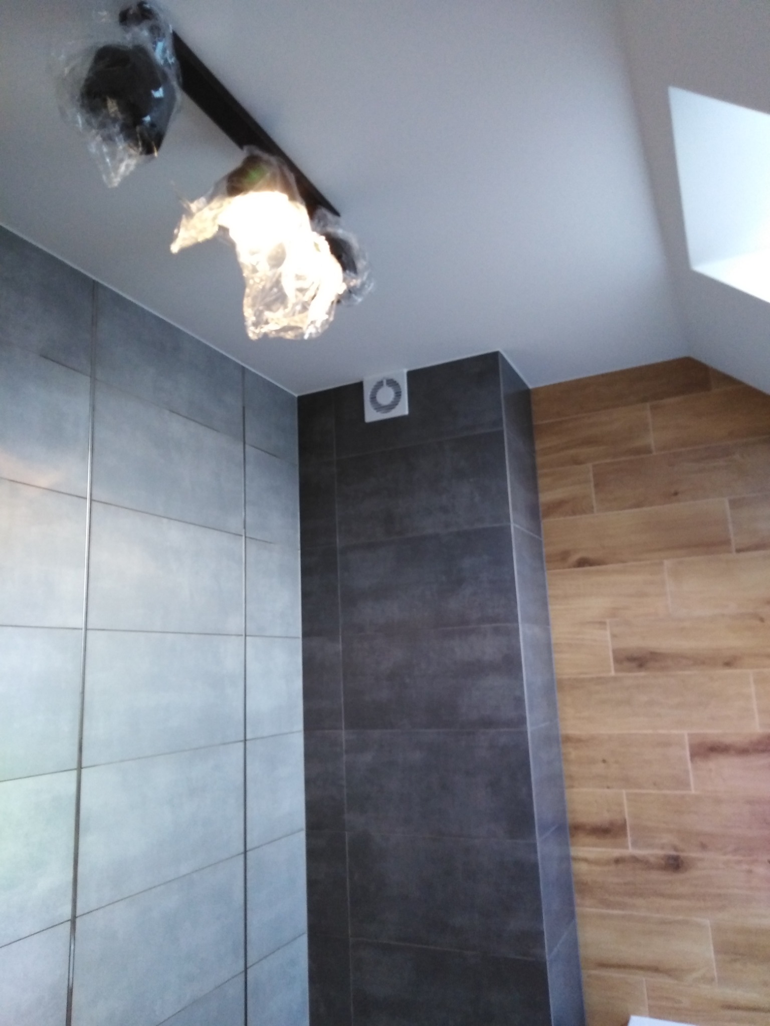 Świeżo wyremontowana łazienka z szarymi i drewnopodobnymi płytkami, oświetleniem sufitowym w folii ochronnej i wentylatorem.