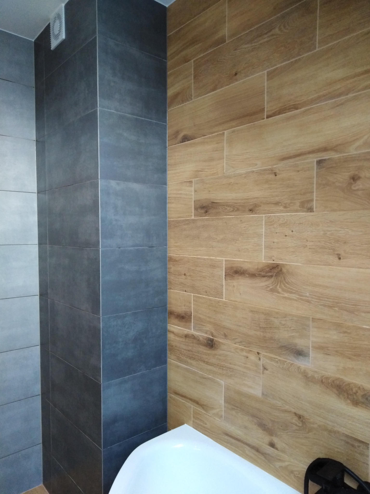 Fragment łazienki z dwoma rodzajami płytek: szare, matowe płytki na ścianie i kolumnie oraz drewnopodobne, podłużne płytki na ścianie obok, widoczny fragment białej wanny.