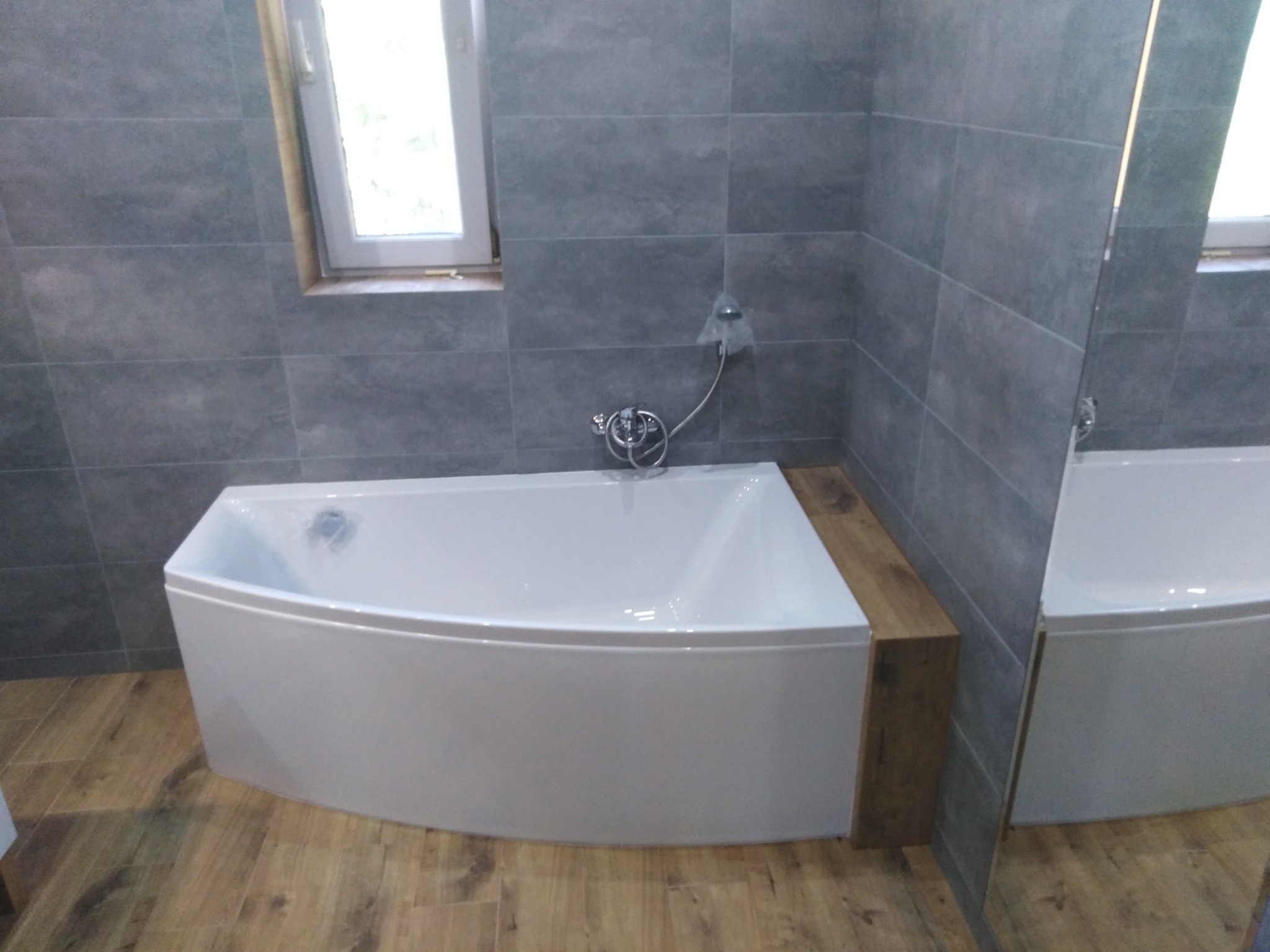 Narożna wanna z baterią i słuchawką prysznicową w nowoczesnej łazience z szarymi płytkami i drewnopodobną podłogą, widoczny fragment lustra.