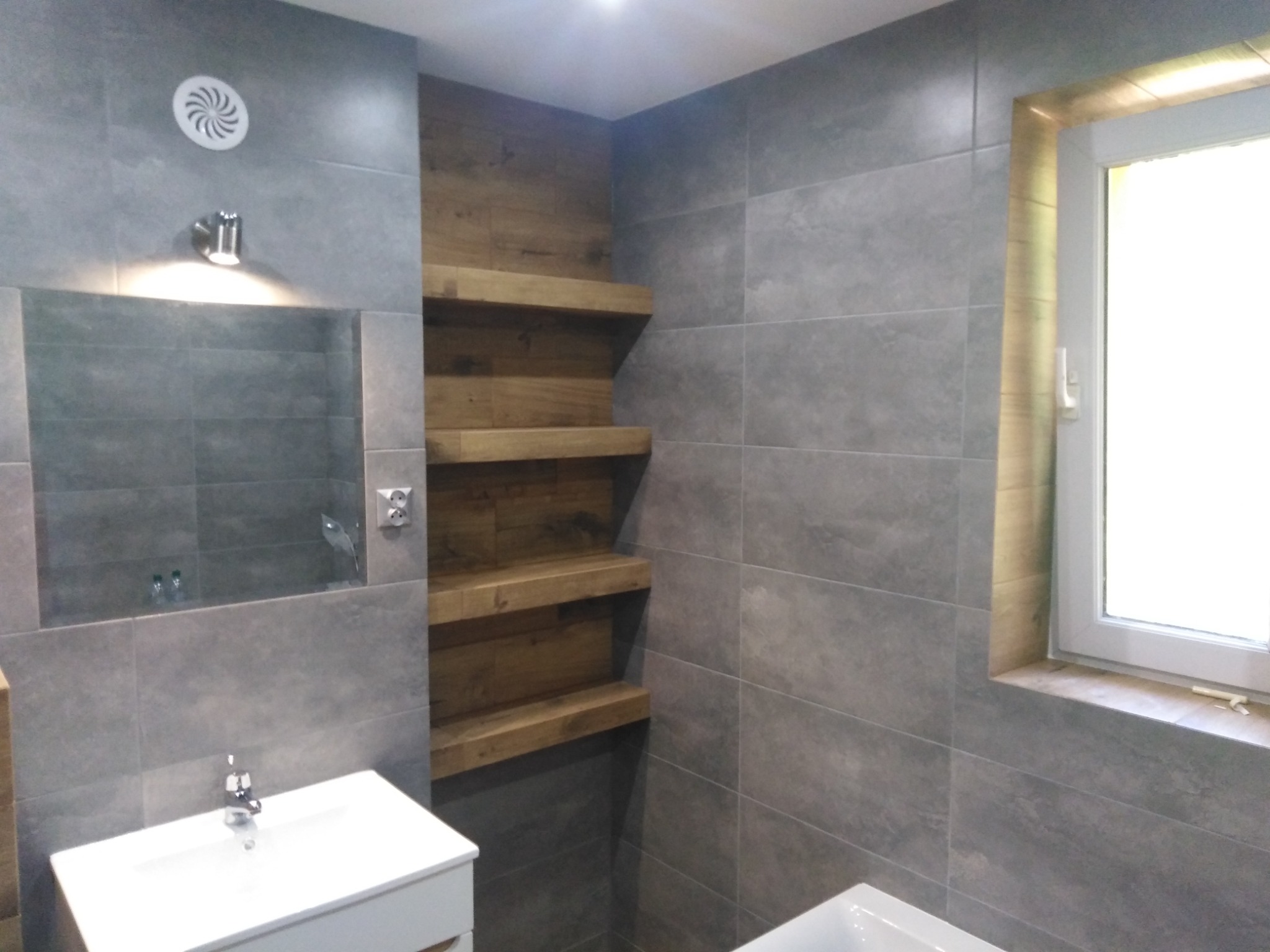 Szara łazienka z drewnianymi półkami wnękowymi, białym zlewem i lustrem, wykończona płytkami imitującymi kamień.