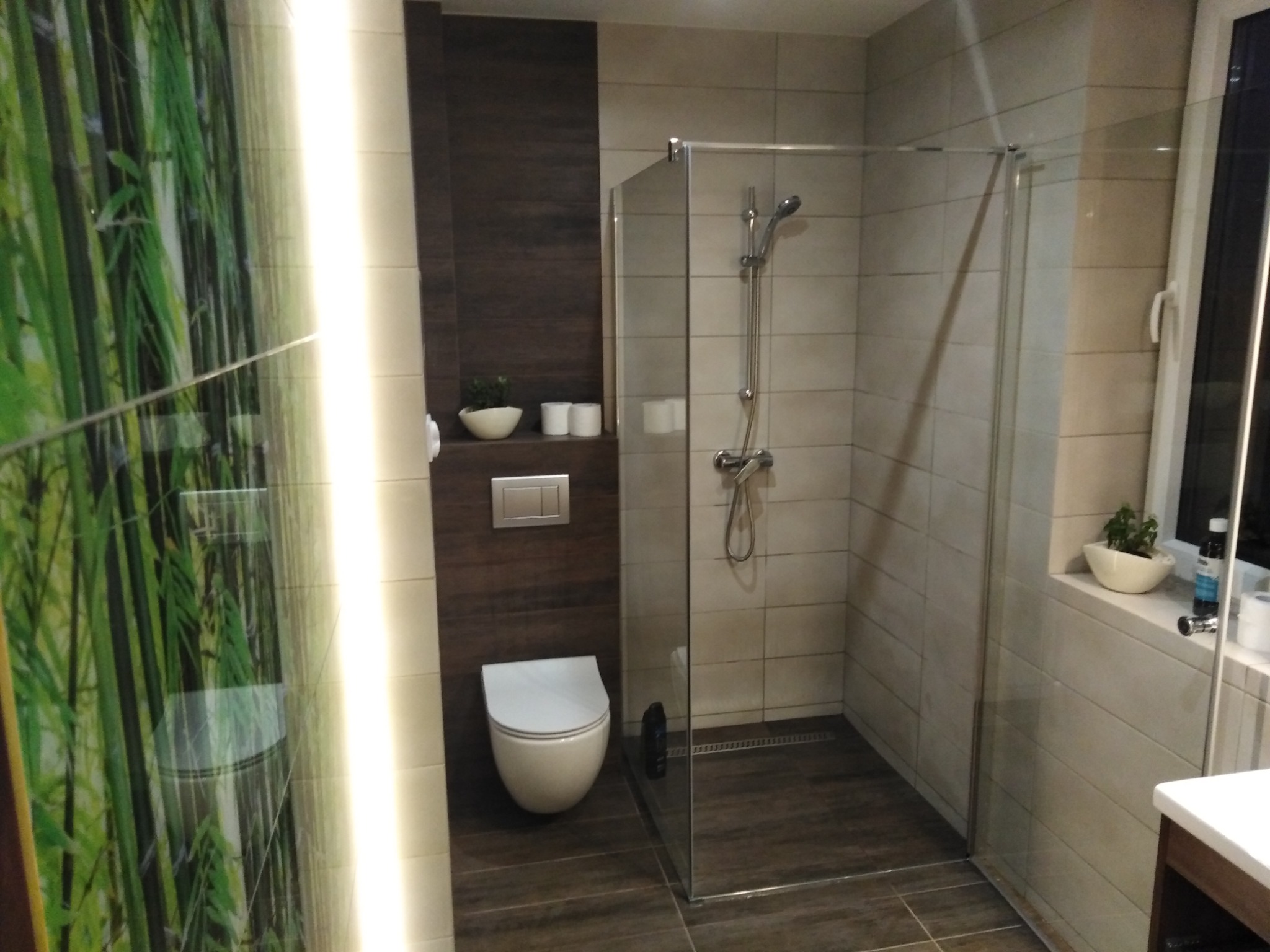 Nowoczesna łazienka z kabiną prysznicową typu walk-in, wiszącą miską WC na tle ciemnobrązowej ściany z drewnopodobnymi płytkami, oraz akcentem zieleni w postaci fototapety z motywem bambusa.
