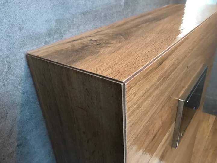 Boczny widok szafki pod umywalkę z frontami imitującymi drewno, z metalowym uchwytem, na tle szarej ściany i drewnianej podłogi.