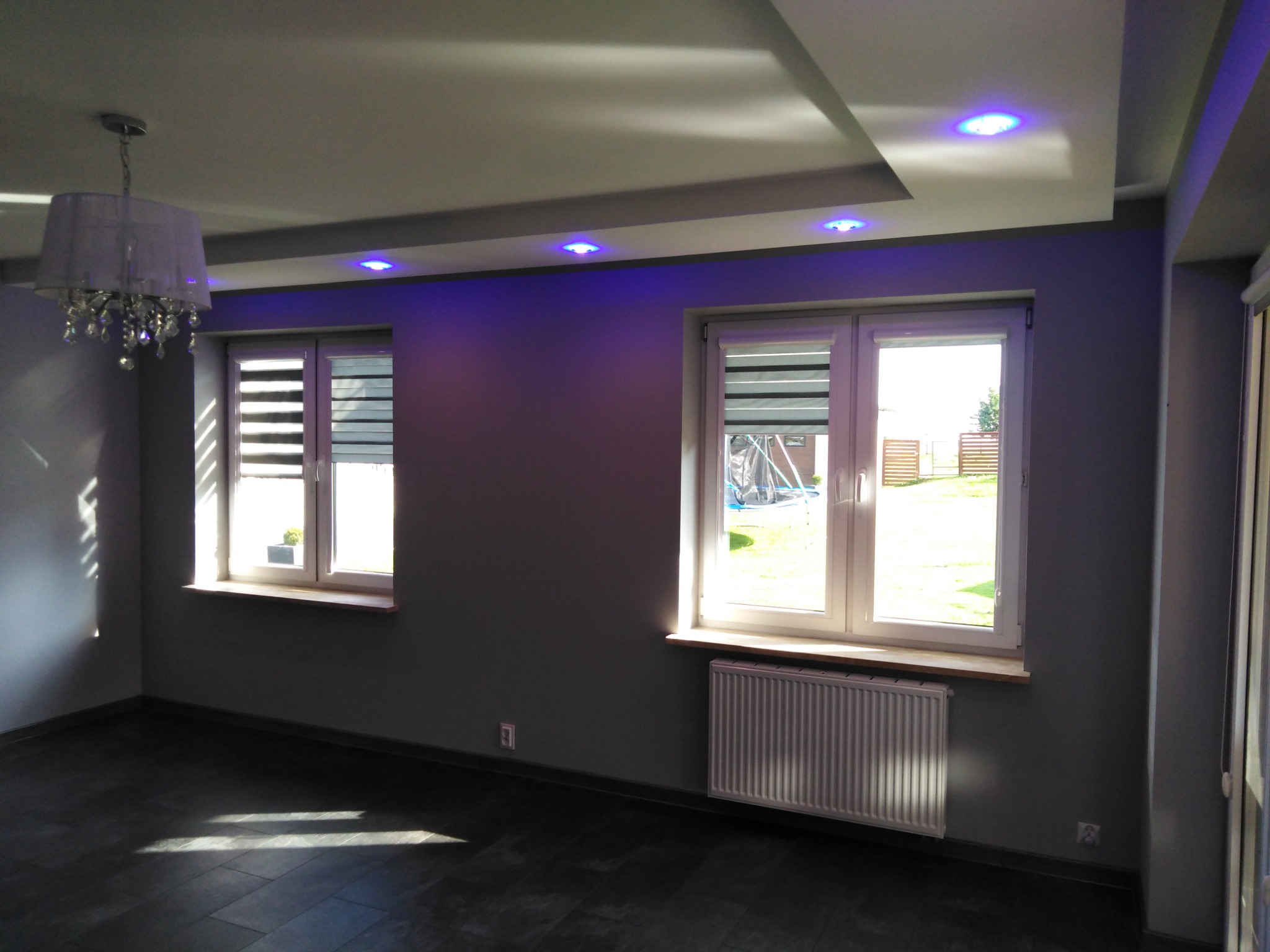 Szare wnętrze pokoju po remoncie z dwoma oknami z żaluzjami, grzejnikiem pod oknem, ozdobnym oświetleniem sufitowym LED w kolorze niebieskim i lampą z kryształkami