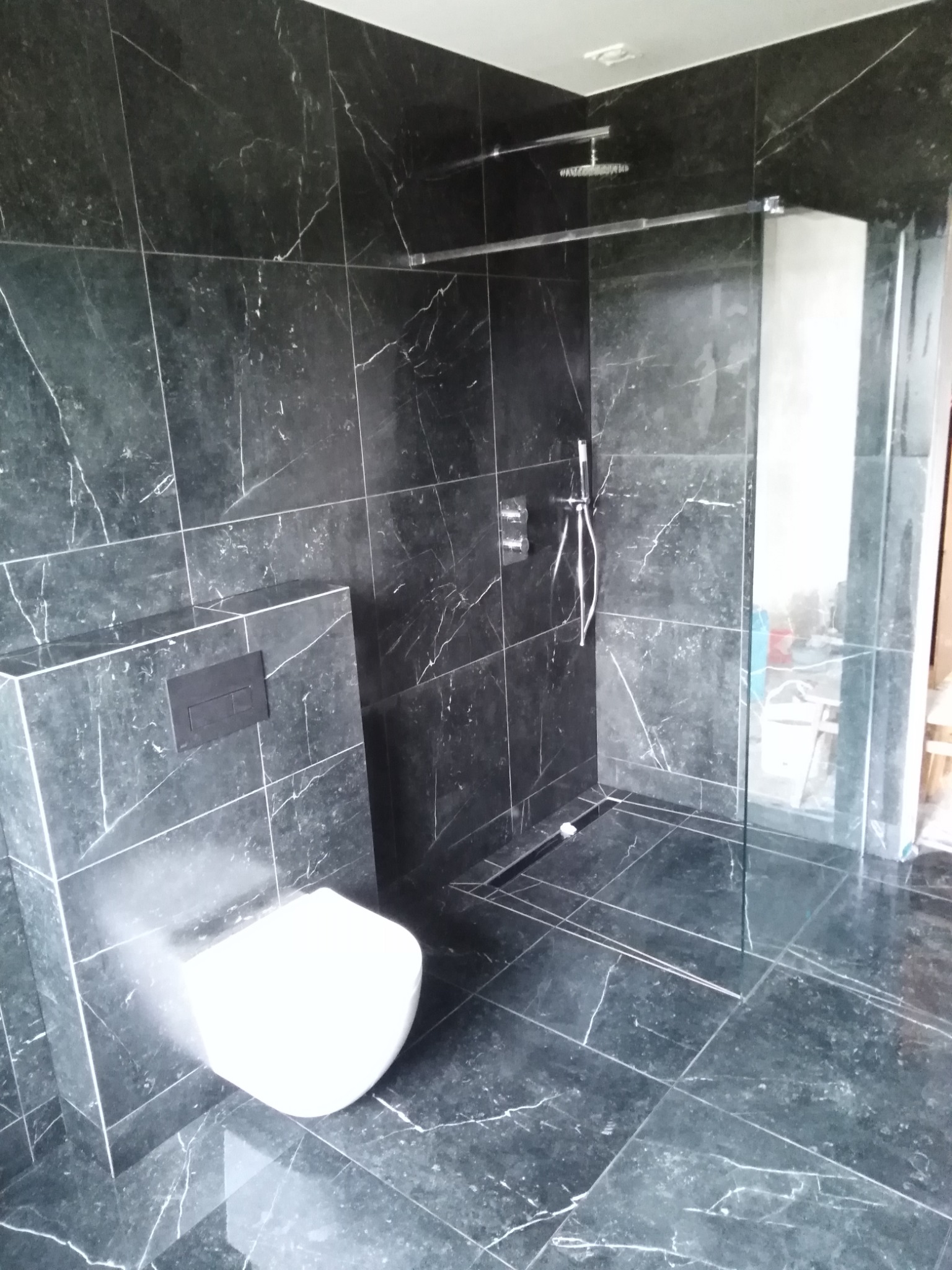 Nowoczesna łazienka wykończona czarnymi płytkami imitującymi marmur z białymi żyłami, z podwieszaną toaletą, szklaną kabiną prysznicową typu walk-in i odpływem liniowym w podłodze.