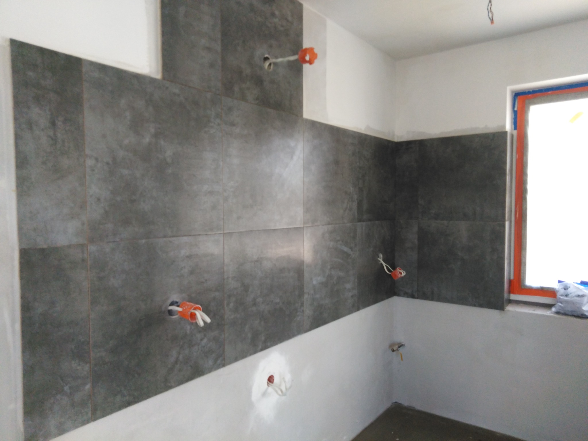 Montaż szarych płytek w łazience, widoczne wystające kable i puszki elektryczne, okno zabezpieczone folią malarską.