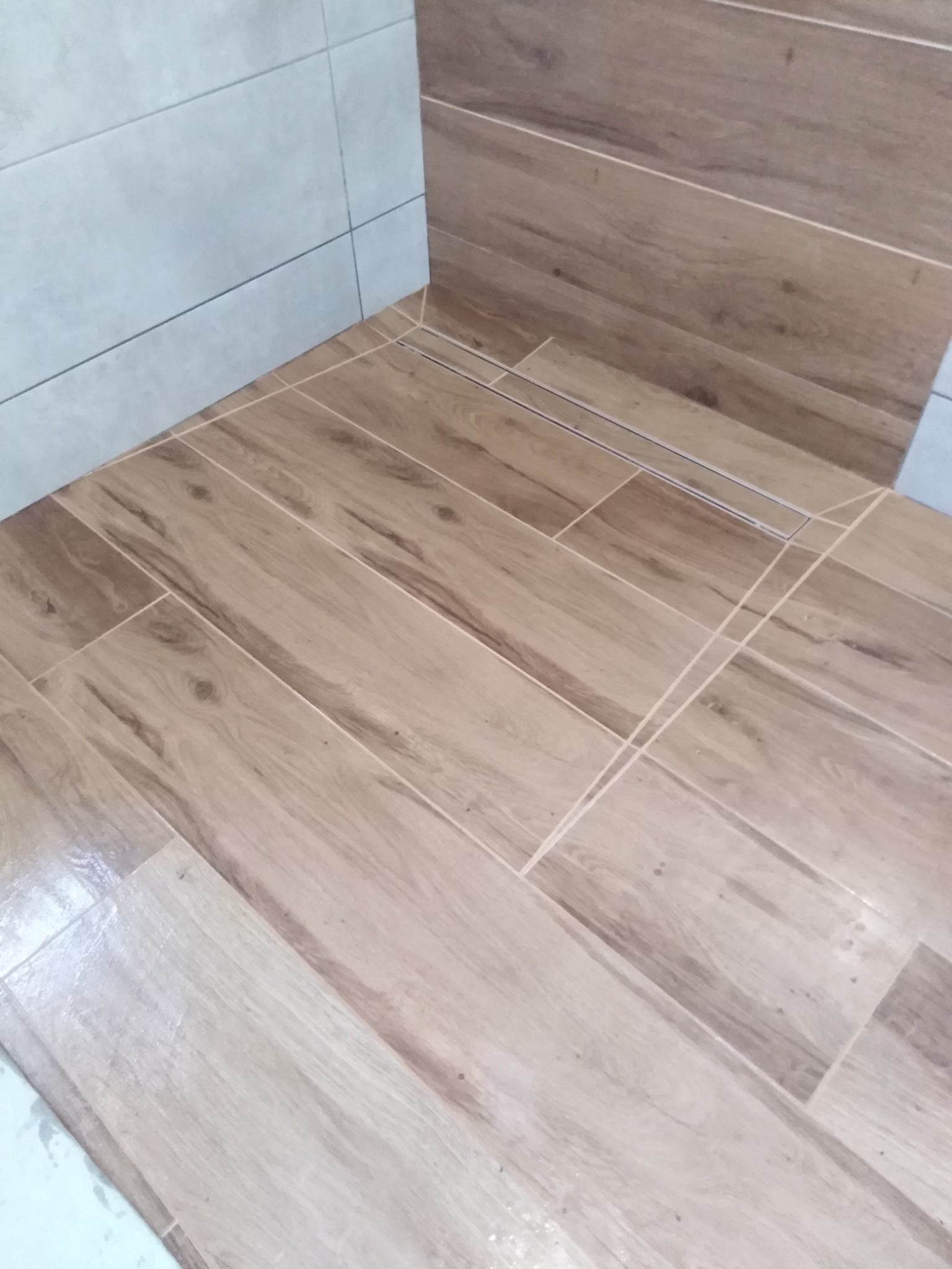 Ułożona podłoga z płytek imitujących drewno w łazience, z widocznym odpływem liniowym w rogu i szarą płytką ścienną u góry.