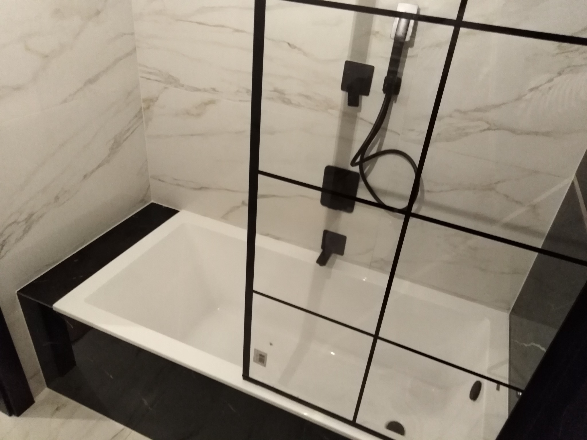 Nowoczesna łazienka z białą wanną obudowaną czarnym marmurem, szklany parawan nawannowy z czarnymi profilami, armatura w kolorze czarnym mat. Ściany wyłożone płytkami imitującymi marmur.
