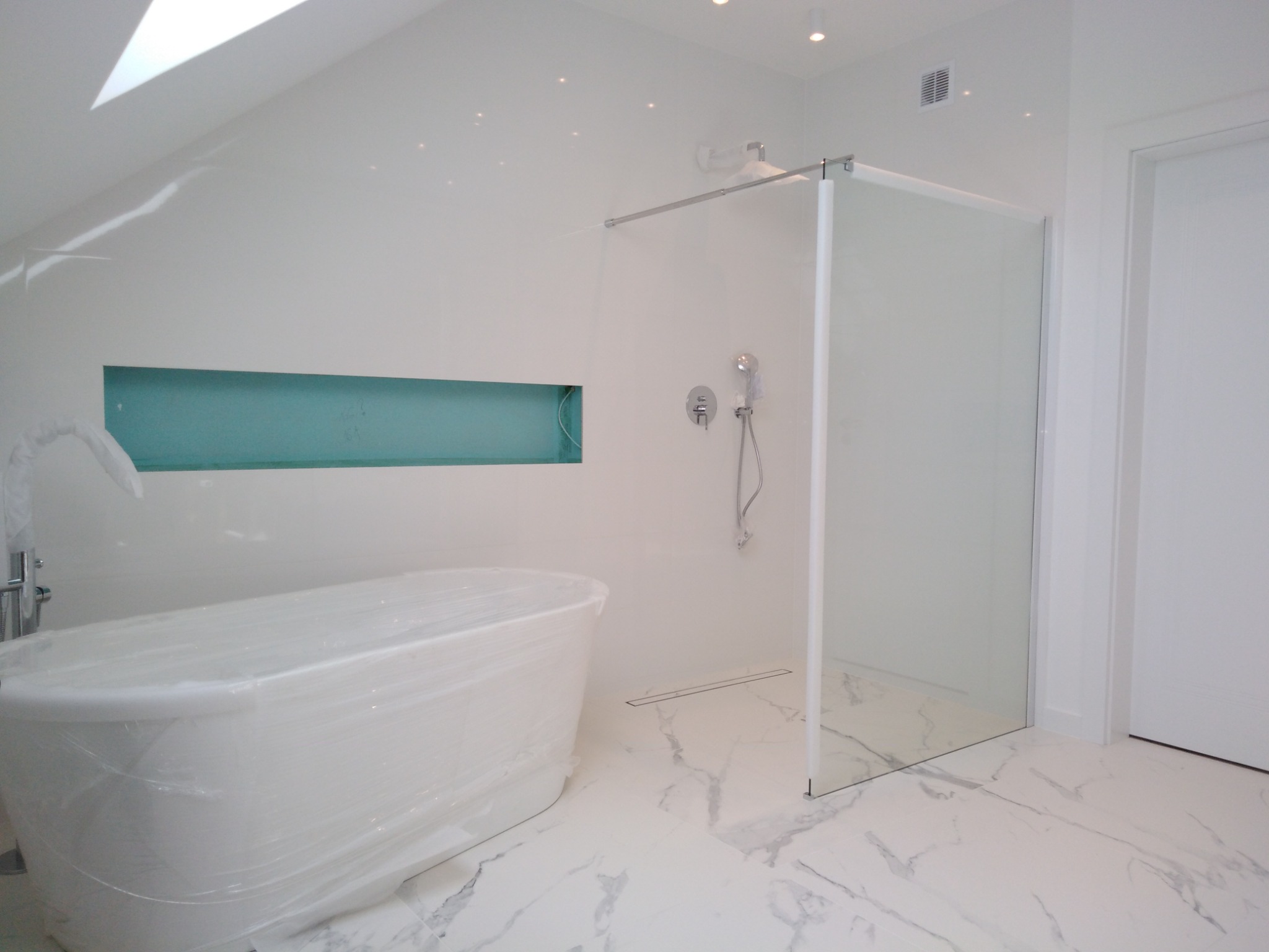 Nowoczesna, jasna łazienka z wolnostojącą wanną owiniętą folią ochronną, szklaną kabiną prysznicową walk-in i marmurową posadzką. W ścianie wnęka z turkusowym tłem. Widoczne punkty świetlne...