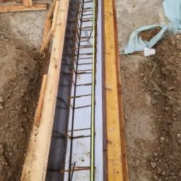 Przygotowanie szalunku i zbrojenia pod wylewkę fundamentu, widoczne drewniane deski, stalowe pręty zbrojeniowe i folia izolacyjna.