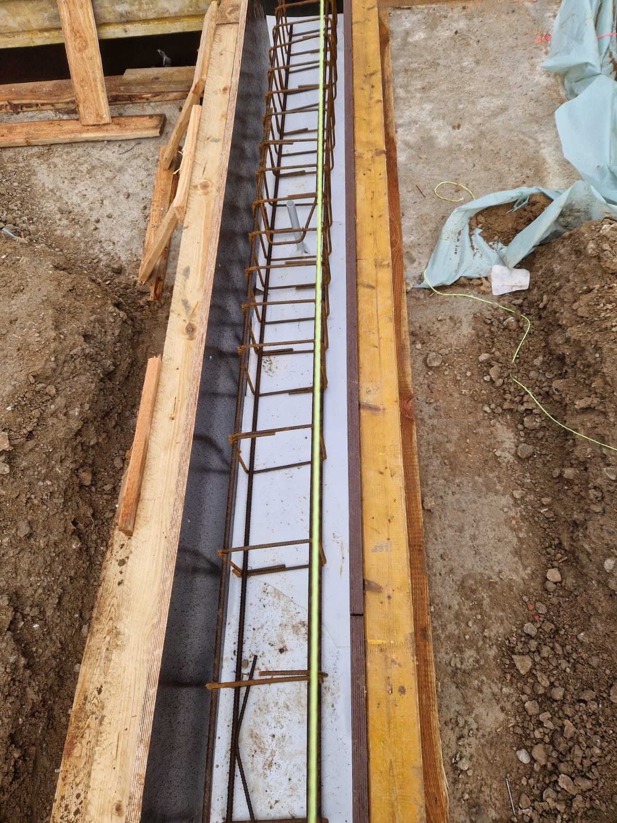 Przygotowanie szalunku i zbrojenia pod wylewkę fundamentu, widoczne drewniane deski, stalowe pręty zbrojeniowe i folia izolacyjna.