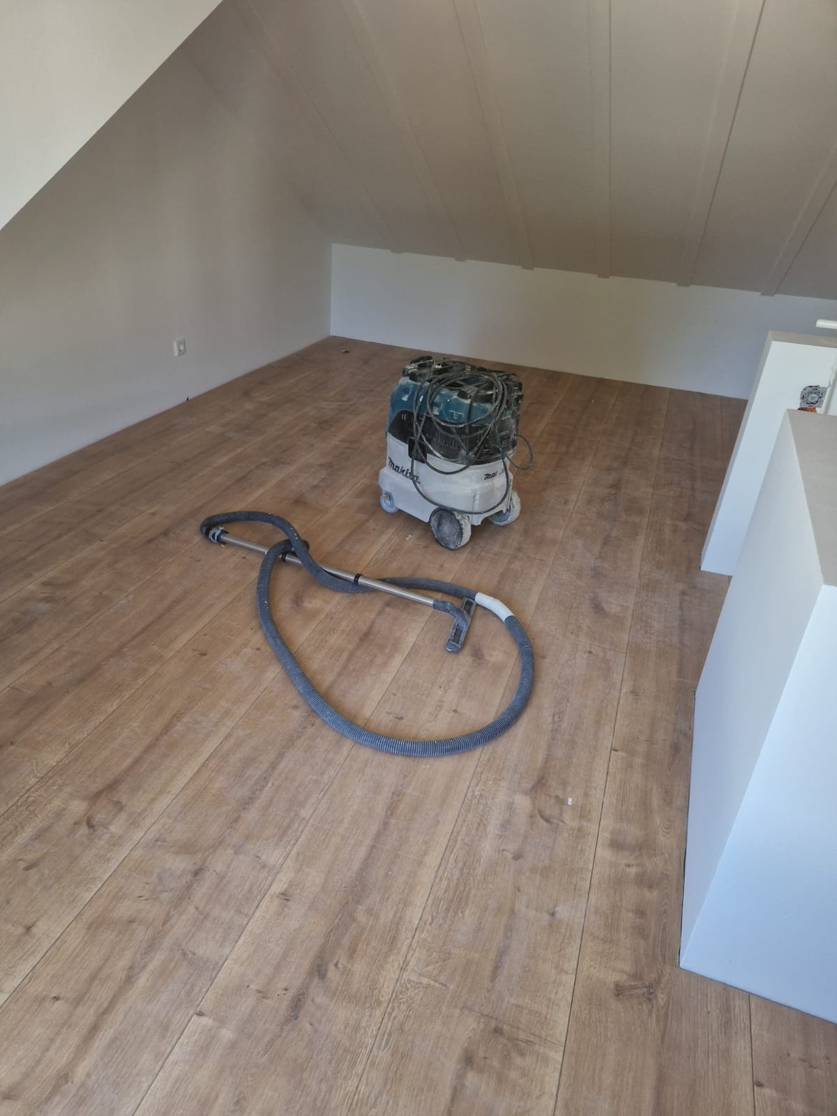Poddasze z drewnopodobną podłogą po remoncie, z widocznym odkurzaczem przemysłowym Makita z wężem na podłodze, ściany pomalowane na biało, skosy sufitowe.