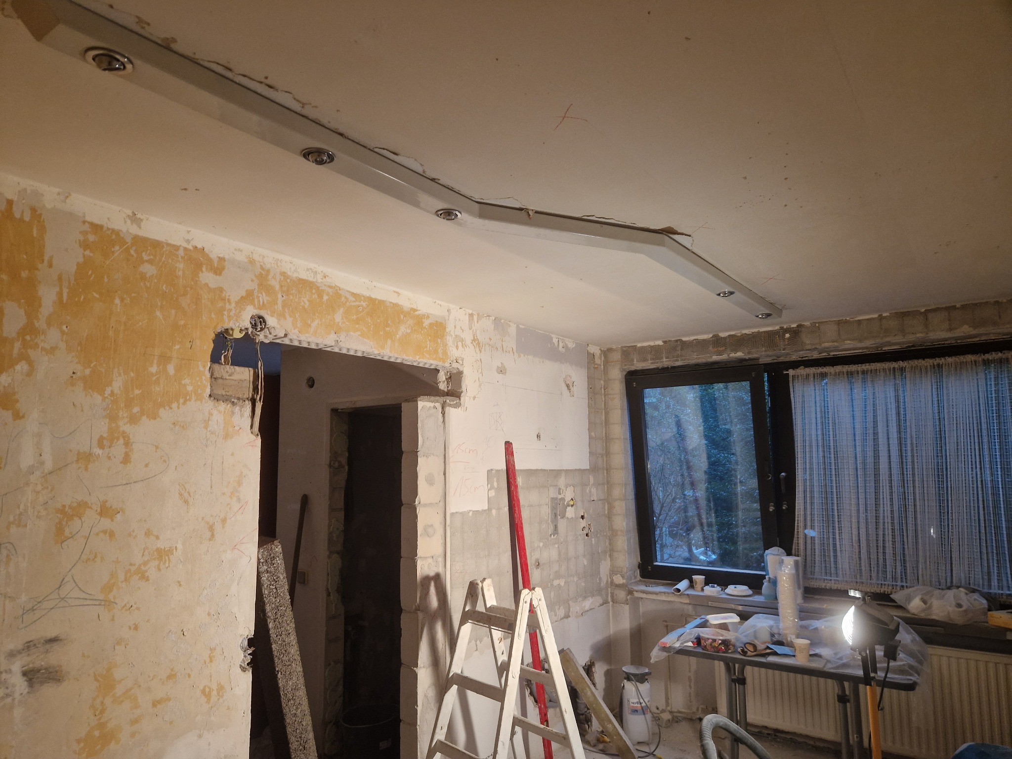 Wnętrze w trakcie remontu z odsłoniętymi fragmentami ścian, drabina oparta o ścianę, widoczne instalacje elektryczne, sufit z zamontowanym oświetleniem szynowym.