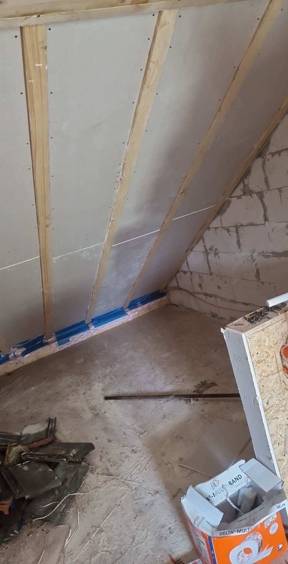 Montaż płyt gipsowo-kartonowych na poddaszu, widoczne drewniane belki konstrukcyjne, niezagruntowane płyty GK, taśma uszczelniająca wzdłuż krawędzi, oraz fragment ściany z bloczków betonowych.