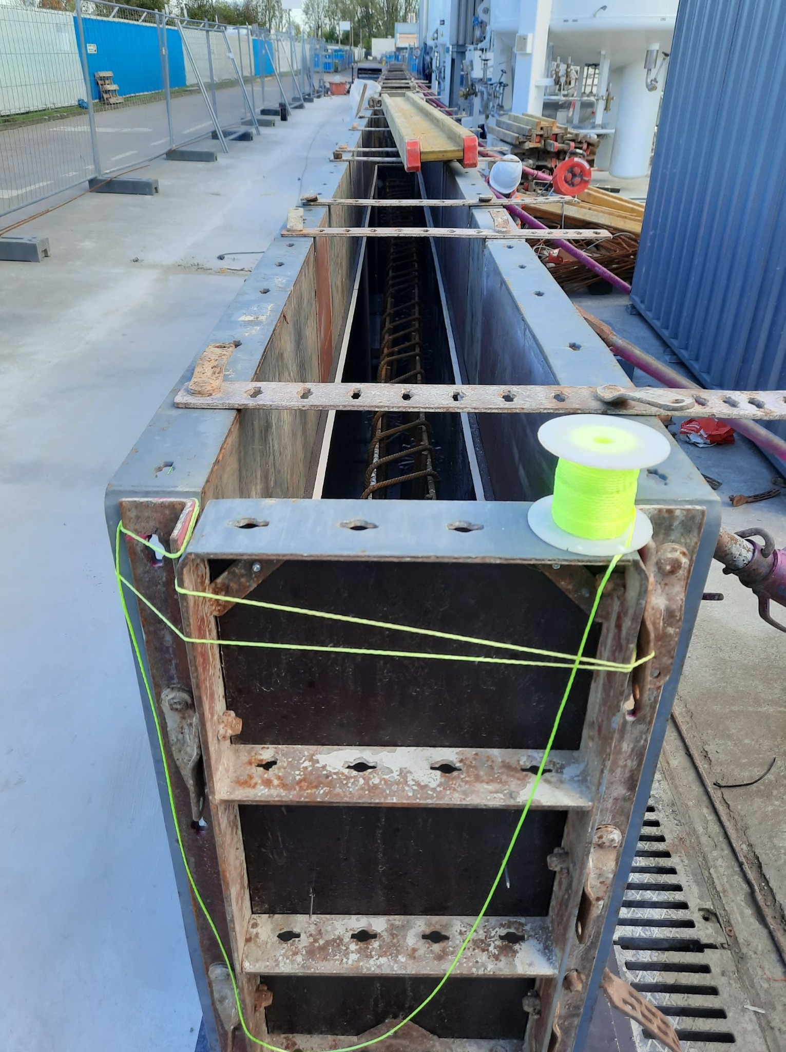 Szczegółowe ujęcie szalunku zbrojeniowego, z neonowo-zieloną linką i szpulą, przygotowanego do zalewania betonem, widoczne stalowe pręty zbrojeniowe wewnątrz, na tle placu budowy.