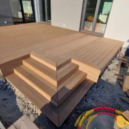 Nowoczesny taras z kompozytowych desek w odcieniach brązu, z czterostopniowymi schodami, widok z góry na plac budowy z elementami wykończeniowymi i zabawkami w tle.