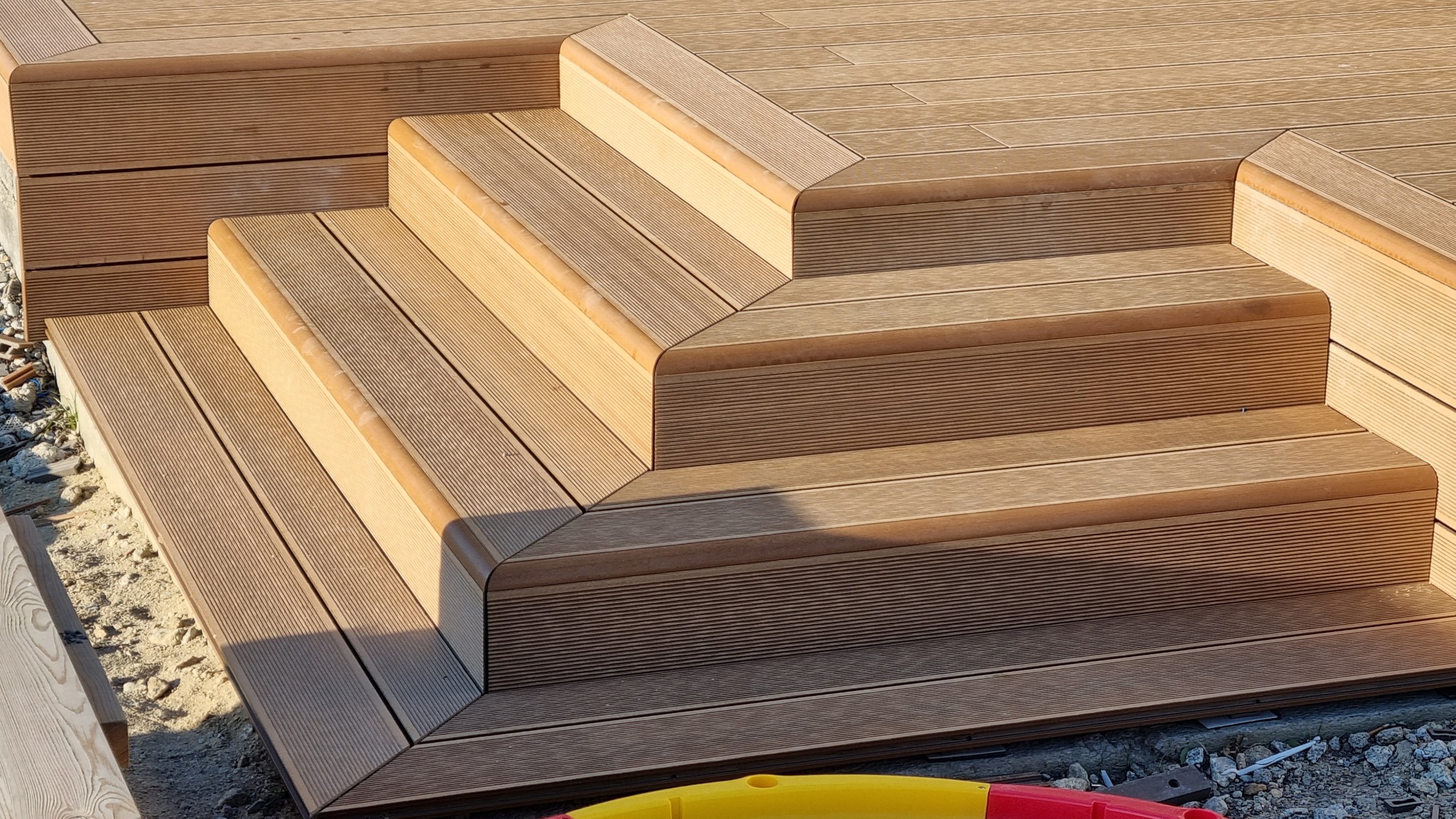 Schody z desek kompozytowych prowadzące na taras, widoczne detale wykończenia krawędzi i połączeń desek, w tle podłoże z gruzem.
