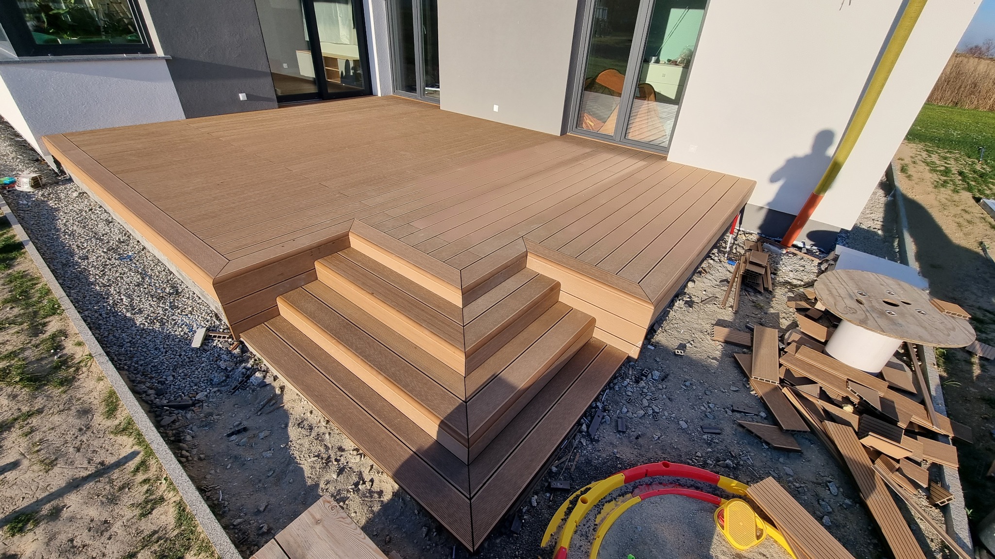 Nowoczesny taras z kompozytowych desek w odcieniach brązu, z czterostopniowymi schodami, widok z góry na plac budowy z elementami wykończeniowymi i zabawkami w tle.