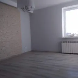 Pusty pokój po remoncie z panelami na podłodze, ścianą wyłożoną dekoracyjną cegłą, grzejnikiem pod oknem i wiszącą lampą.