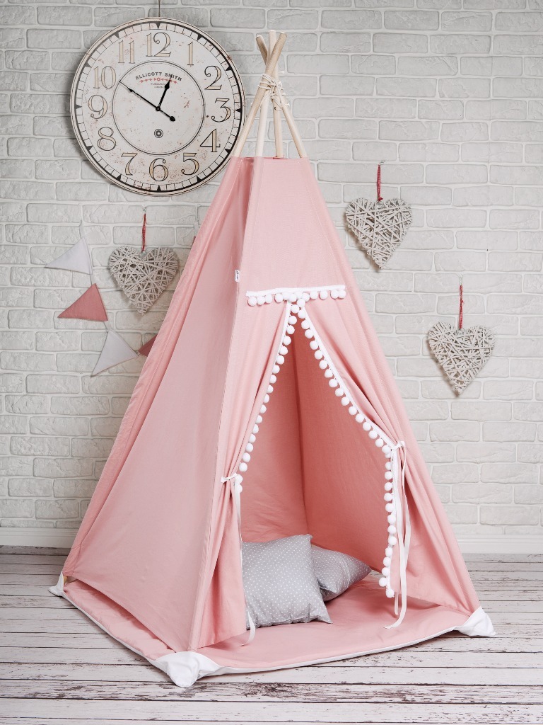 Różowe tipi dla dzieci z białymi pomponami i szarymi poduszkami w środku, ustawione na drewnianej podłodze przed ceglaną ścianą, ozdobione wiszącymi sercami i zegarem.
